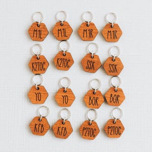 May include: A collection of sixteen wooden keychains. Each keychain is a hexagon shape with a silver ring. The keychains are engraved with various three-letter and four-letter combinations in a dark color.