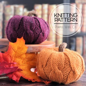 May include: Two knitted pumpkins, one deep purple and one orange, with brown stems, sit on a wooden surface. Autumn leaves in shades of red, orange, and yellow are in the foreground. The image includes text that reads "Knitting Pattern" and "Purple Wool Co."