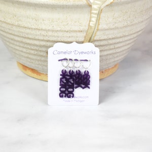 May include: Set of four purple stitch markers with the words "Sock", "Heel", "Toe", and "K2tog" on them. The markers are attached to silver rings and are displayed on a white card with the text "Camelot DyeWorks" and "Made in Michigan".