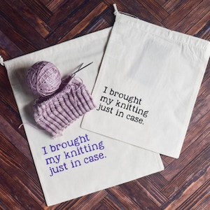 May include: Two off-white canvas bags with drawstring closures, each printed with the text "I brought my knitting just in case." A ball of lavender yarn and a partially knitted piece are next to one bag. The background is a wooden surface.