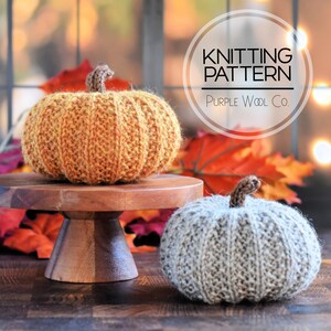 May include: Two knitted pumpkins, one orange and one gray, are displayed. The orange pumpkin sits on a wooden pedestal. The pumpkins have brown stems. The image includes text that reads "Knitting Pattern" and "Purple Wool Co."