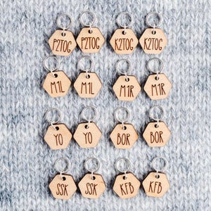 May include: A set of 16 wooden knitting stitch markers with different knitting abbreviations engraved on them. The markers are hexagonal in shape and have silver metal rings.