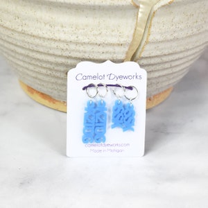 May include: Two blue stitch markers with the words "Cozy" and "Bossy" on them. The markers are attached to silver rings and are hanging from a white card with the text "Camelot Dyeworks" and "Made in Michigan".