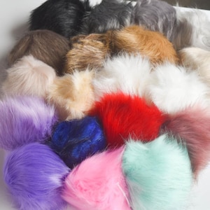 May include: A collection of fluffy faux fur pom-poms in various colors, including black, gray, brown, white, red, pink, purple, blue, and mint green. The pom-poms are densely packed together, showcasing their soft texture and vibrant hues. Ideal for craft projects.