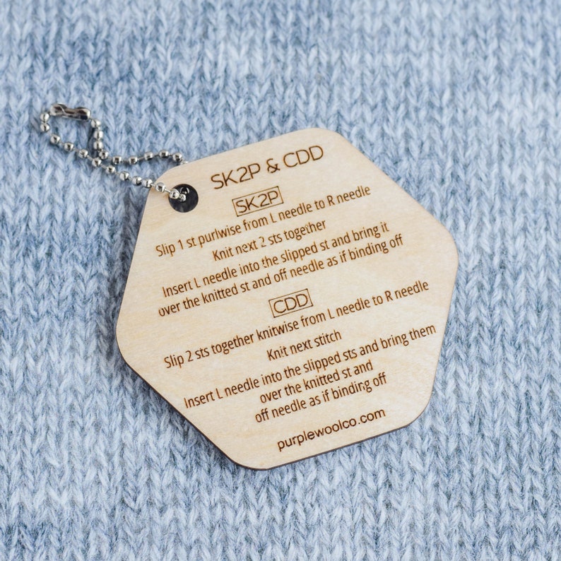 Puede incluir: Un marcador de punto de tejido de madera con una cadena plateada, con instrucciones para las t&eacute;cnicas de tejido SK2P y CDD. El marcador es de color marr&oacute;n claro con texto negro y est&aacute; sobre un fondo de tejido azul claro.
