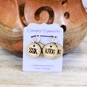 May include: Three wooden stitch markers with silver rings and black lettering. The markers read "SSK", "K2TOG", and "DC". The markers are on a purple and white card that reads "Camelot DyeWorks" and "Made in Michigan".