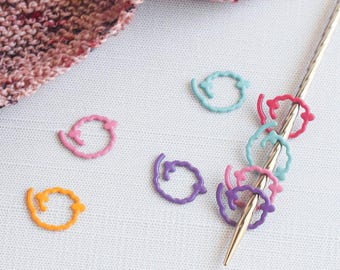 May include: A collection of colorful stitch markers in various shades of pink, purple, blue, and orange. Some markers are on a gold knitting needle, while others are scattered on a white surface. A knitted piece in shades of pink and red is in the background.
