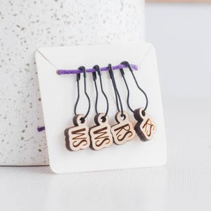 May include: A set of four wooden stitch markers with black metal clasps, attached to a white card with a purple string. Each marker has a different letter combination: WS, WS, RS, and RS. The markers are made of light-colored wood.
