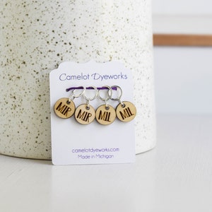 May include: A set of four wooden stitch markers with silver rings, attached to a white card. Each marker is round and engraved with the letters "MIR" or "MIL". The card reads "Camelot Dyeworks" and "Made in Michigan".