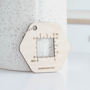 May include: Wooden gauge with a square window for measuring yarn or fabric. It has markings in inches and centimeters. The gauge is attached to a chain and has the text "purplewoolco.com" on the bottom.