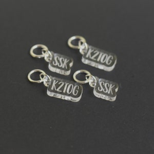 May include: Four clear acrylic stitch markers with silver rings. The markers are labeled "K2tog", "SSK", and "K2tog" and "SSK".