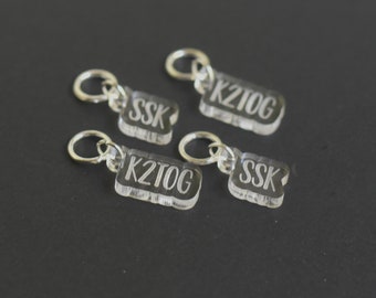 May include: Four clear acrylic stitch markers with silver rings. The markers are labeled "K2tog", "SSK", and "K2tog" and "SSK".