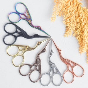 May include: A collection of decorative stork-shaped scissors in various metallic finishes, including rainbow, black, gold, copper, silver, and rose gold. The scissors are arranged in a circular pattern, showcasing their intricate designs and sharp blades. Ideal for crafting and sewing.