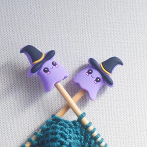 May include: Two purple ghost-shaped knitting needle toppers with black witch hats and smiling faces. The needles are crossed over teal yarn, with a light gray background. The toppers add a festive touch to knitting projects.