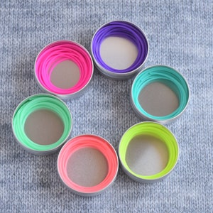 May include: Six round containers with lids, each containing a different color of elastic string. The containers are arranged in a circle on a gray knitted fabric.