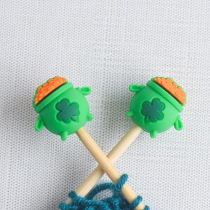 May include: Two wooden knitting needles with green pot of gold toppers, each featuring a shamrock. The pots have orange 'gold' and are crossed over blue yarn, set against a white background.