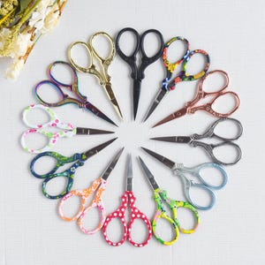 May include: A collection of decorative embroidery scissors arranged in a circle. The scissors feature various colors and patterns, including floral designs, gold, black, and iridescent finishes. The blades are silver-toned, and the handles have ornate details.