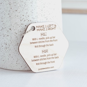 May include: Wooden knitting stitch marker with a chain attached. The marker is hexagonal and has the text "MAKE 1 LEFT & MAKE 1 RIGHT" on the top. The marker also has instructions for "M1L" and "M1R" stitches.