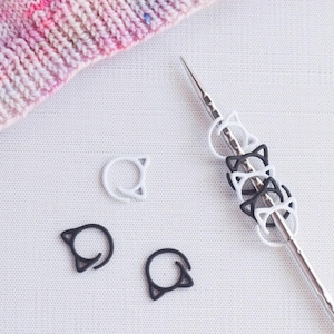 May include: A close-up of a set of white and black cat-shaped stitch markers. The markers are strung on a silver knitting needle.