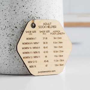 May include: Wooden hexagonal guide for determining sock size based on shoe size. The guide features a chart with women's and men's shoe sizes in US and EU sizing, along with the corresponding approximate foot length in inches and centimeters. The guide is labeled "Adult Sock Helper" and includes the website address "purplewoolco.com".