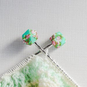 May include: Two silver knitting needles with floral patterned green and white tips. The needles are crossed and resting on a white and green knitted fabric.