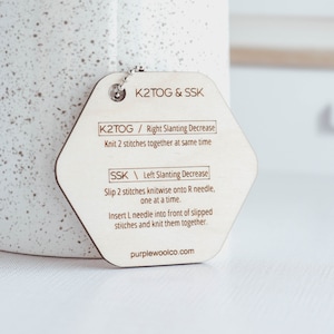 May include: A wooden hexagonal tag with instructions for knitting techniques, "K2TOG & SSK". The tag has the text "K2TOG / Right Slanting Decrease Knit 2 stitches together at same time" and "SSK / Left Slanting Decrease Slip 2 stitches knitwise onto R needle, one at a time. Insert L needle into front of slipped stitches and knit them together." The tag also has the website "purplewoolco.com".