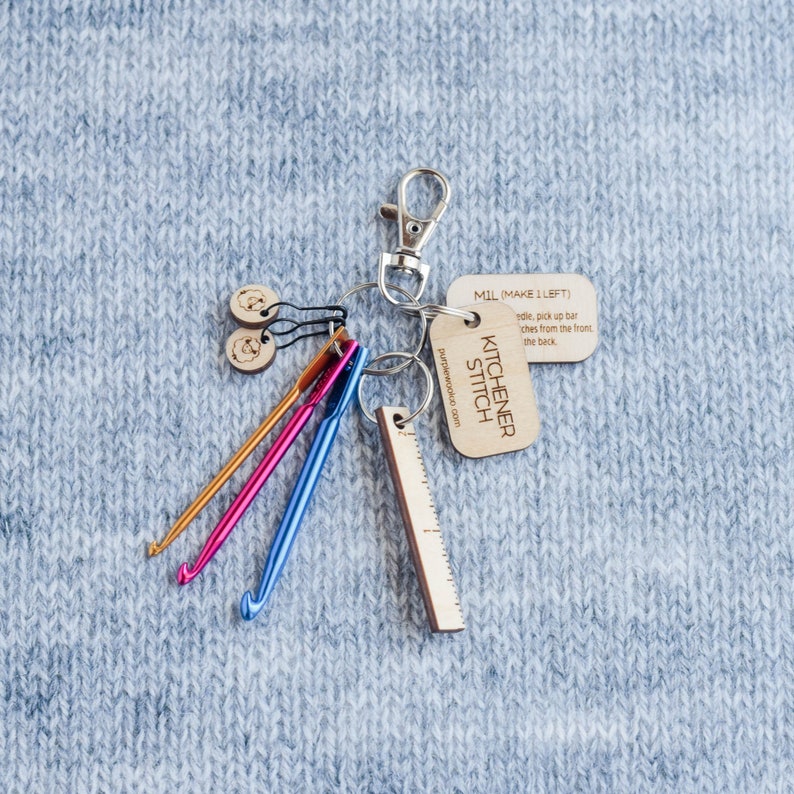 May include: A set of knitting tools on a keychain, including a ruler, crochet hooks, and wooden tags that say "KITCHENER STITCH" and "MIL (MAKE 1 LEFT)".