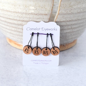 May include: Four wooden stitch markers on a white card. Each marker is a small, round wooden disc with black text. The card reads "Camelot Dyeworks" and "Made in Michigan". The markers are connected by a purple thread.