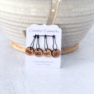 May include: A set of four wooden stitch markers with the words "FRONT", "BACK", "RIGHT", and "LEFT" printed on them. The markers are attached to black loops and a purple bar. The card reads "Camelot Dyeworks" and "camelotdyeworks.com Made in Michigan".