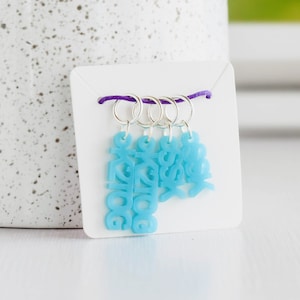 May include: Set of four blue stitch markers with the words "Dog", "Cat", "Fish", and "Bird" on them. The markers are attached to a purple cord.