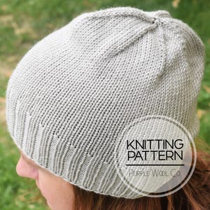 May include: A light gray knitted hat with a ribbed brim. The hat is made of a soft, textured yarn and has a rounded crown. The text "KNITTING PATTERN" and "Purple Wool Co." is visible on the hat.