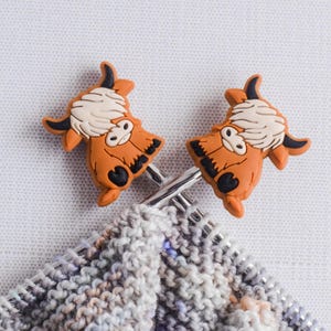 May include: Two brown and white cow-shaped knitting needle stoppers on a pair of knitting needles with a grey and white knitted fabric.