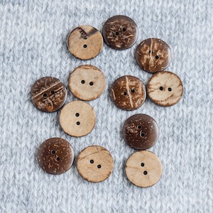 May include: A collection of 11 brown coconut shell buttons with two holes each. The buttons are arranged in a semi-circle on a gray knitted fabric background.