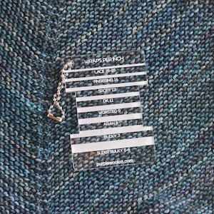 May include: A clear plastic gauge with a silver chain attached. The gauge has a white background with black text that reads "Wraps Per Inch" and lists different yarn weights with corresponding wrap counts per inch. The text at the bottom reads "purplewoolco.com".