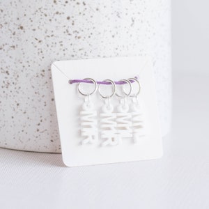 May include: Four white stitch markers with the word "OMR" on each. The markers are attached to a purple string and are displayed on a white card.