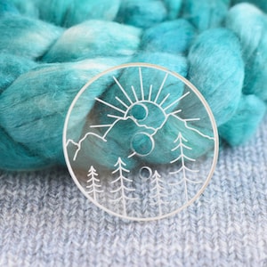 May include: A clear acrylic yarn bowl with a white design of a mountain range, sun, and moon. The bowl is sitting on a gray knitted fabric.