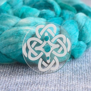 May include: A clear glass yarn bowl with a white Celtic knot design. The bowl is sitting on a gray knitted surface.