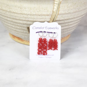 May include: Three red stitch markers with the word "GOOG" written on them. The markers are attached to silver rings and are displayed on a white card with the text "Camelot Dyeworks" and "Made in Michigan".