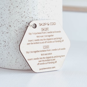 Puede incluir: Una guía de puntos de tejido de madera con el texto "SK2P & CDD" e instrucciones para técnicas de tejido. La etiqueta es hexagonal con una pequeña cadena de metal adjunta. El texto "purplewoolco.com" está impreso en la parte inferior.