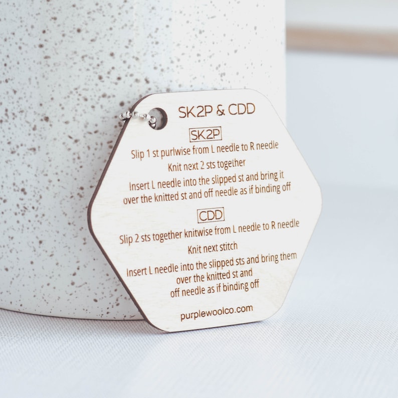 Puede incluir: Una gu&iacute;a de puntos de tejido de madera con el texto "SK2P & CDD" e instrucciones para t&eacute;cnicas de tejido. La etiqueta es hexagonal con una peque&ntilde;a cadena de metal adjunta. El texto "purplewoolco.com" est&aacute; impreso en la parte inferior.