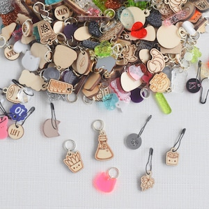 May include: A variety of colorful and wooden stitch markers, including a pink heart, a green leaf, a cat, a bottle, a crown, a french fry, and a leaf. The markers are scattered on a white surface.