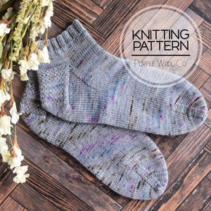 May include: A pair of grey knit ankle socks featuring a speckled pattern with small flecks of purple, blue, and brown. The socks have a ribbed cuff and a rounded toe. Text overlay reads KNITTING PATTERN Purple Wool Co.