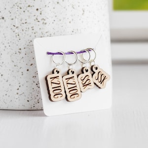 May include: Four wooden stitch markers with purple string. The markers are labeled "K2tog", "K2tog", "SSK", and "SSK".