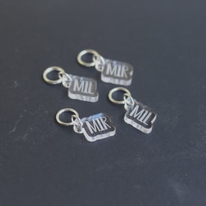 May include: Four clear acrylic stitch markers with silver rings. The markers are engraved with the letters "MIL" and "MIR".