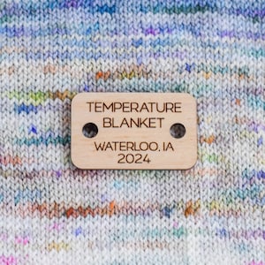 May include: A wooden tag with the text "TEMPERATURE BLANKET WATERLOO, IA 2024" on a colorful knitted blanket.