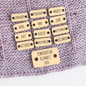 May include: A collection of wooden tags with months of the year and the text "TEMPERATURE BLANKET 2026" printed on them. The tags are arranged on a light purple knitted blanket.