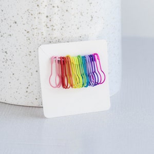May include: A rainbow assortment of safety pins on a white card. The pins are arranged in a row, with the colors transitioning from pink to purple.