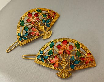 May include: Two gold-colored fan-shaped hair clips. Each clip features a floral design with red, orange, blue, and green enamel. The intricate cut-out design and the U-shaped pin add to the clip's decorative appeal.