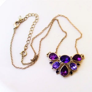 May include: A gold-toned necklace featuring a pendant with a cluster of purple and violet gemstones. The gemstones are in various shapes, including oval and teardrop, and are set in a gold-colored metal.