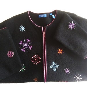 May include: Black cardigan with a pink zipper and colorful snowflake embroidery. The cardigan has a pink trim around the neckline and cuffs.